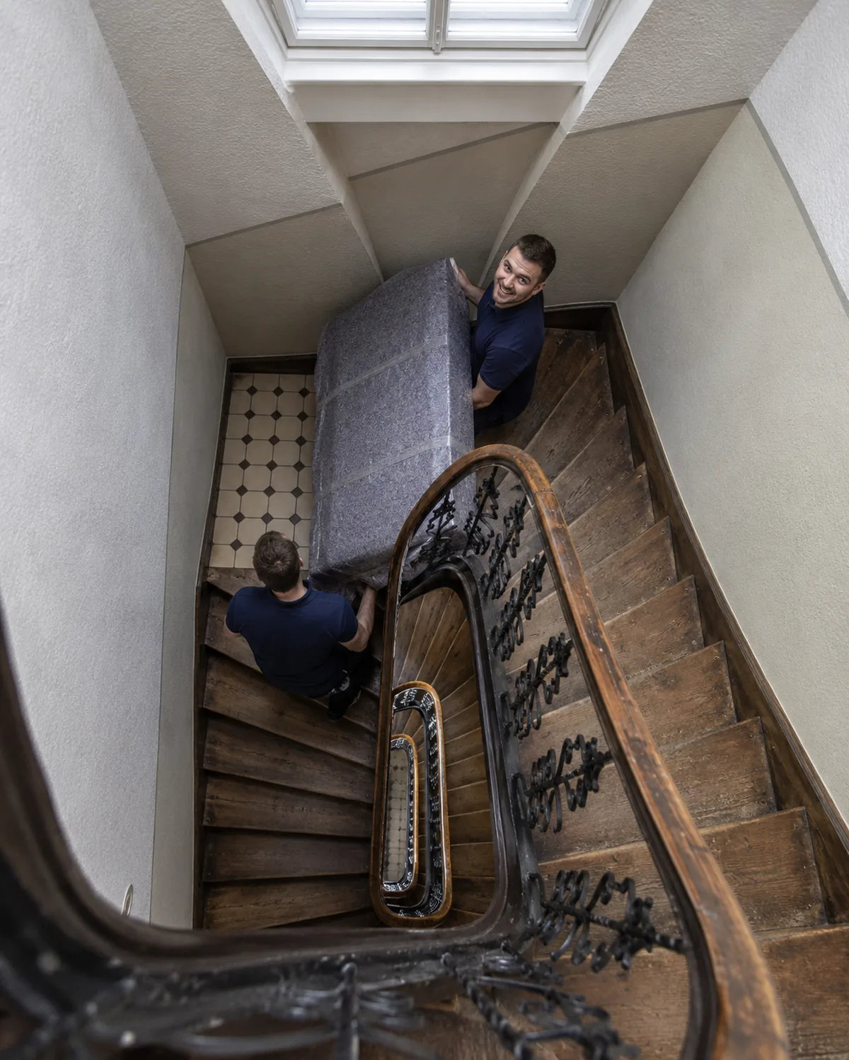 Blick von oben in ein enges Bremer Treppenhaus, zwei Umzugshelfer tragen eine verpackte Matratze um die Wendung